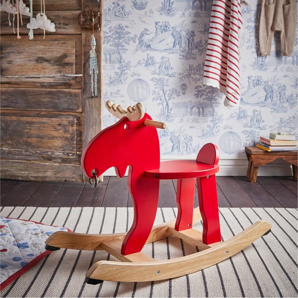 Un élan à bascule en rouge et en hévéa sur un tapis rayé blanc et noir avec une couverture matelassée blanche/rouge/bleue sur le côté.