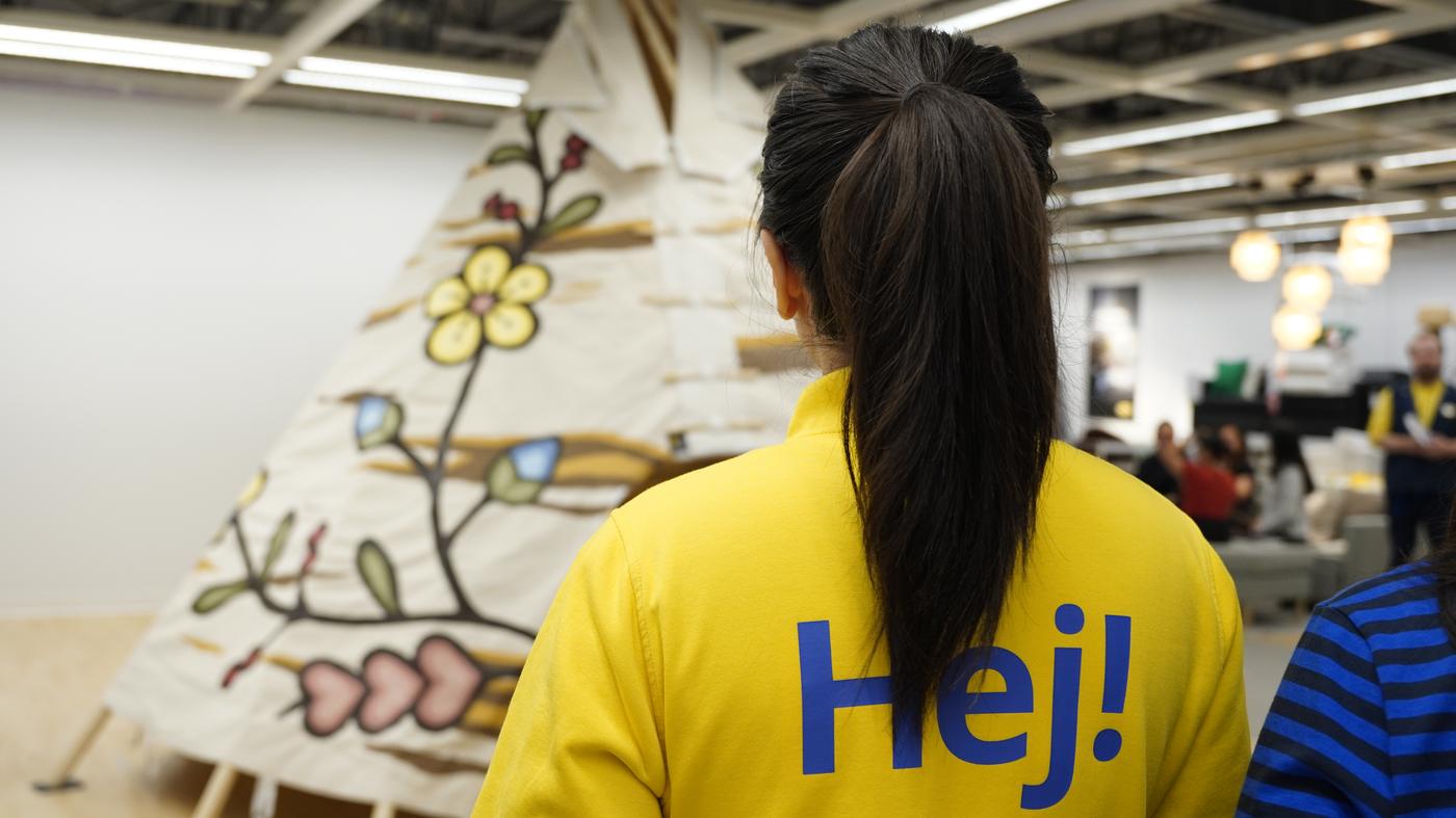 Un employé d'IKEA, vêtu d'une veste jaune portant l'inscription « HEJ ! » en bleu dans le dos, se tient debout en regardant un tipi dans un magasin IKEA.