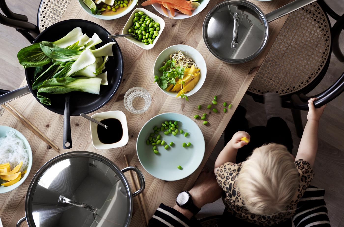 Un enfant assis sur les genoux d’un adulte mange avec les doigts à une table en bois où des fruits et des légumes tranchés sont posés dans des bols et des casseroles.