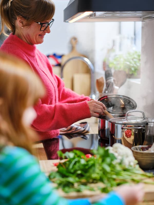 Un enfant devant un plan de travail tandis qu’une adulte en pull rouge remue dans une casserole IKEA 365+ en acier inoxydable à l’arrière d’une plaque à induction.