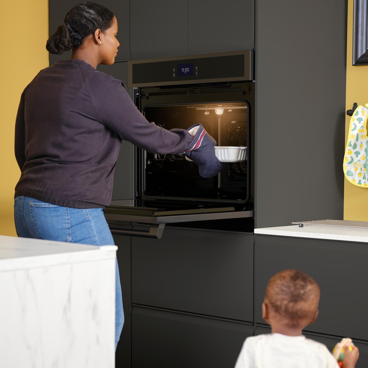 Un enfant en bas âge regarde une femme sortir un plat d’un four mural HJÄLTEBY dans une cuisine aux façades anthracite mat.