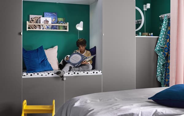 Un enfant lit dans ce petit coin douillet aménagé entre deux rangements gris et comportant coussins, lampe et minibibliothèque.