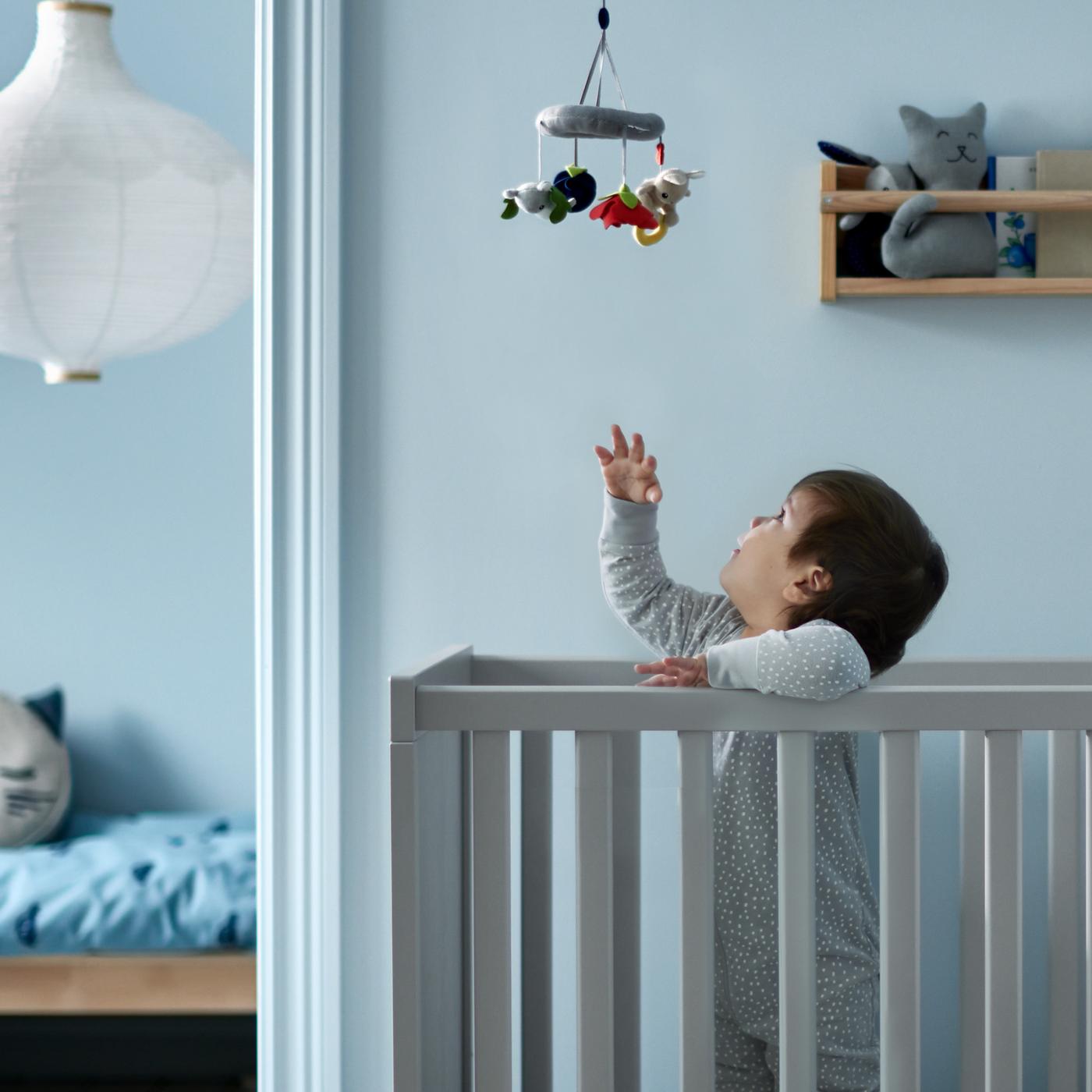 Un enfant se tient debout dans un lit bébé SUNDVIK gris et tend la main vers le mobile multicolore GULLIGAST suspendu au-dessus de lui.