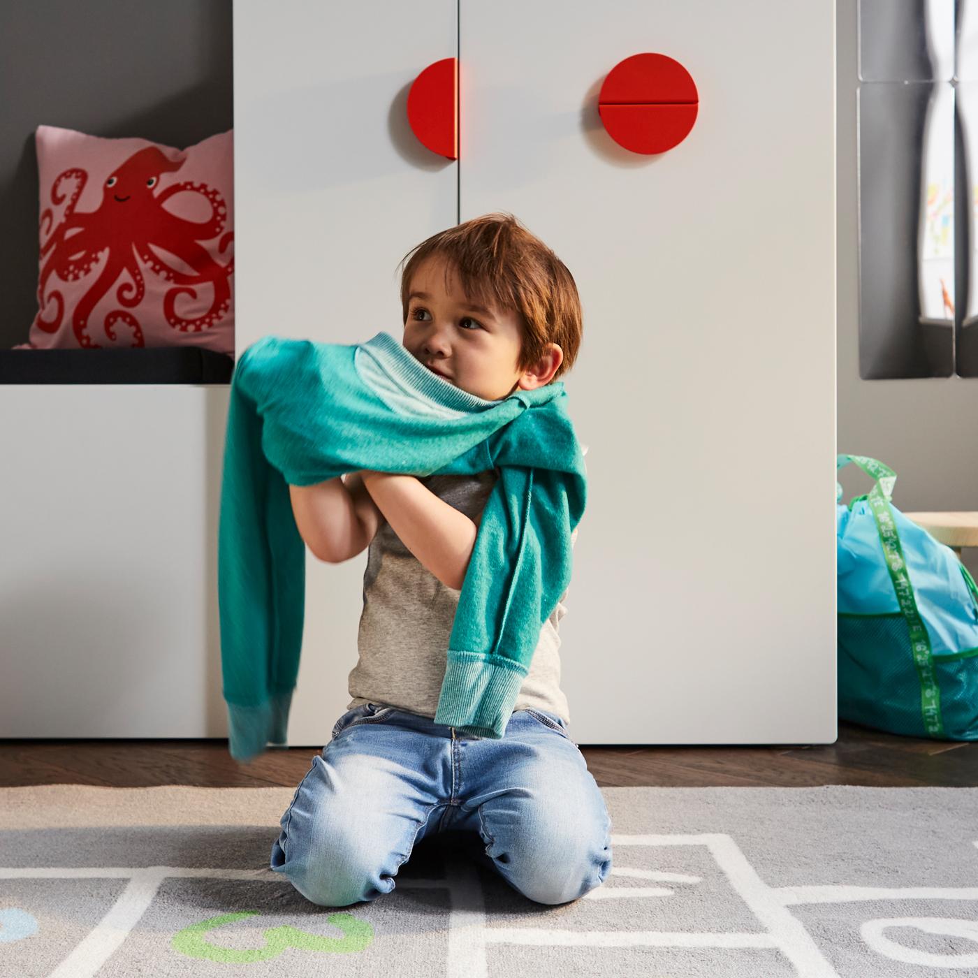Un enfant s’habille sur un tapis gris devant un banc blanc avec rangement pour jouets et une armoire blanche avec rangement coulissant.