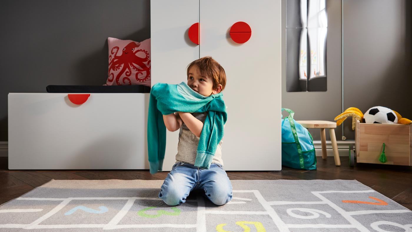 Un enfant s’habille sur un tapis gris devant un banc blanc avec rangement pour jouets et une armoire blanche avec rangement coulissant.