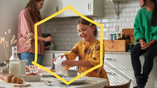 Un enfant vêtu d’une robe jaune met une serviette dans un verre sur une table, tandis qu’une autre personne dans une cuisine lumineuse avec des carreaux blancs remue des ingrédients dans un bol.