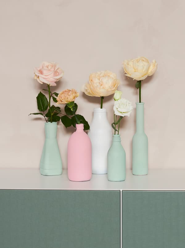 Un ensemble de vieilles bouteilles de différentes formes peintes dans des tons pastel et transformées en vases contenant chacun une fleur SMYCKA.