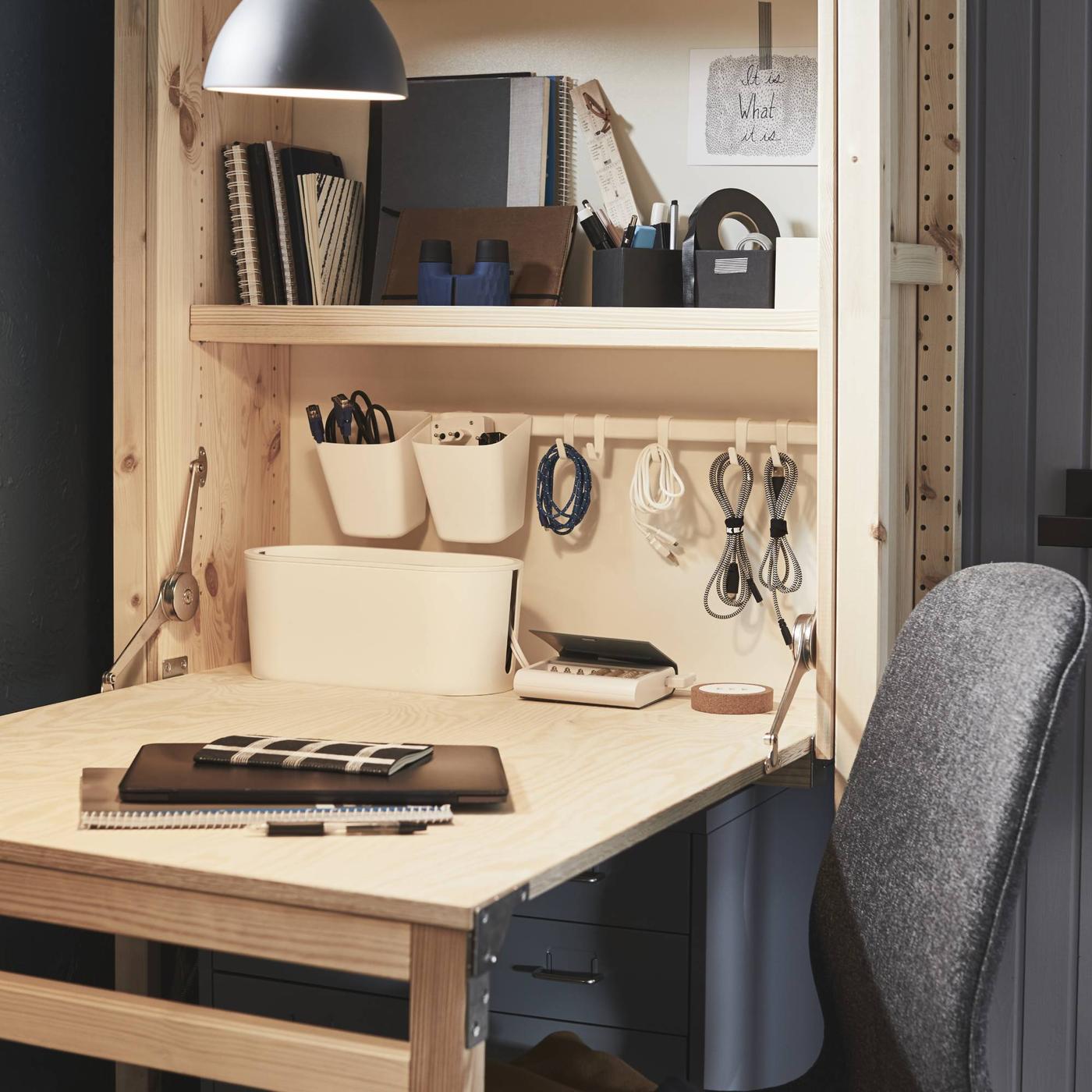 Un escritorio plegable IVAR de madera de pino, con accesorios de escritorio en un estante lateral y sobre la mesa, frente a una silla de oficina gris.