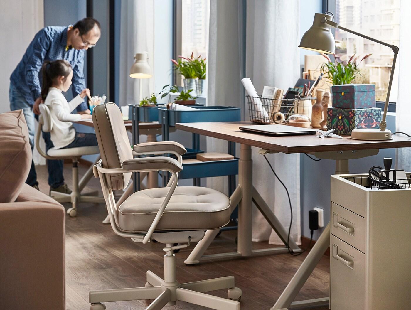 Un espace de bureau à domicile avec un bureau pour un adulte et un enfant, avec un père et une fille en arrière-plan.