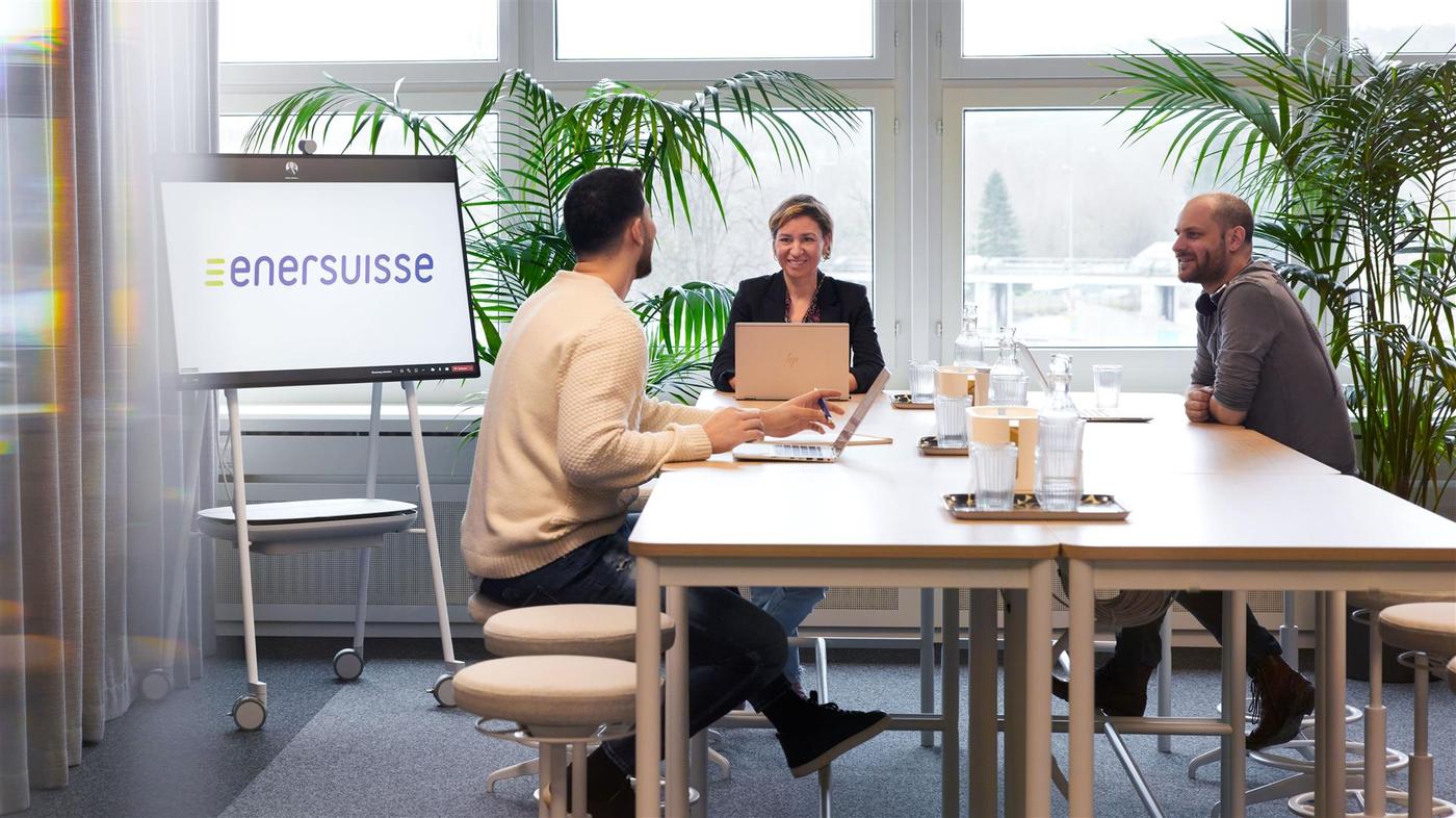 Un espace pour les présentations et les réunions, avec des bureaux blancs permettant de s’asseoir ou de se tenir debout. Trois collaborateur-trice-s discutent. 