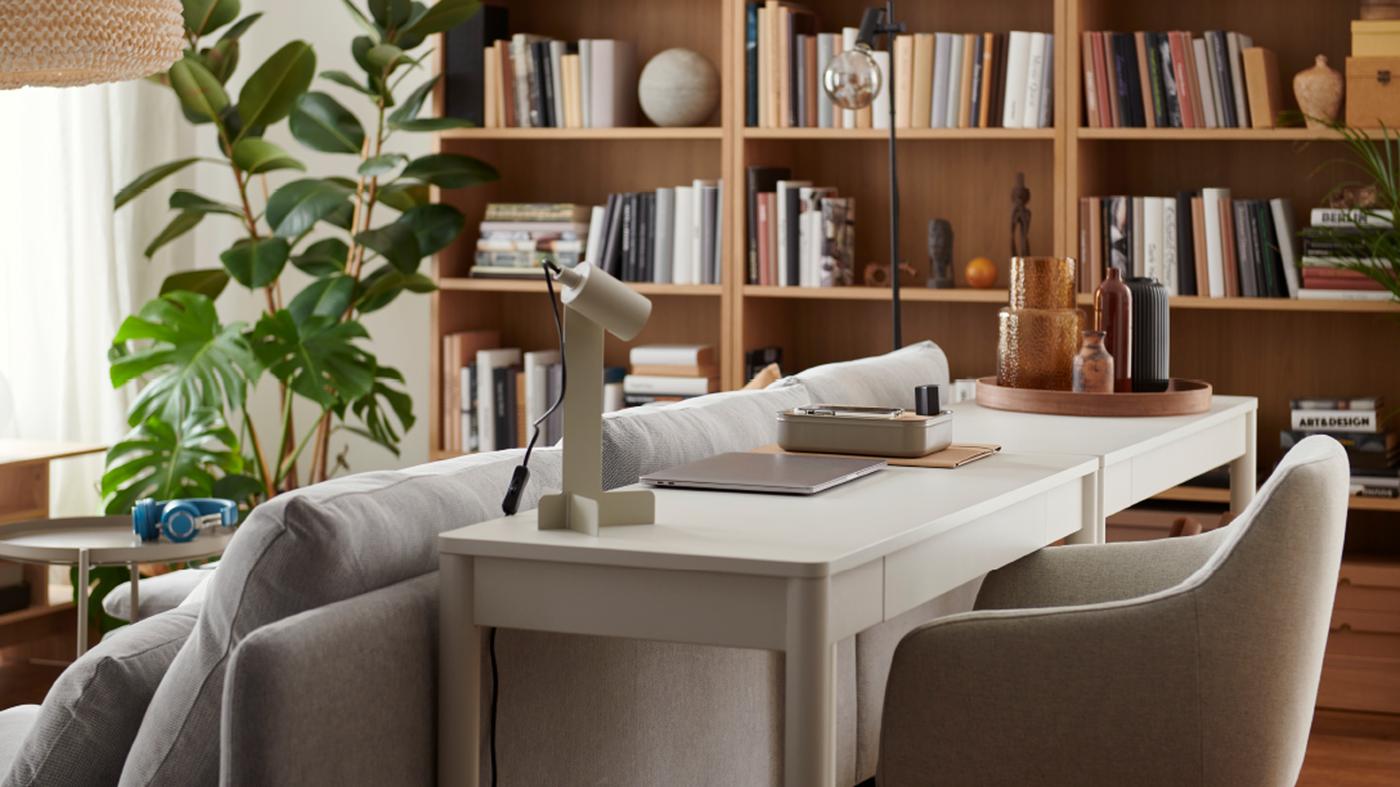 Un espace salon avec un petit poste de travail installé derrière un canapé et composé d’un bureau TONSTAD blanc et d’une lampe FLOTTILJ.
