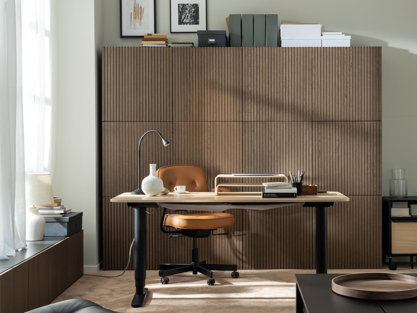 Un espacio de trabajo iluminado por el sol con un escritorio para trabajar de pie/sentado BEKANT, una silla de oficina ALEFJÄLL y una pared de gabinetes BESTÅ con frentes de madera.