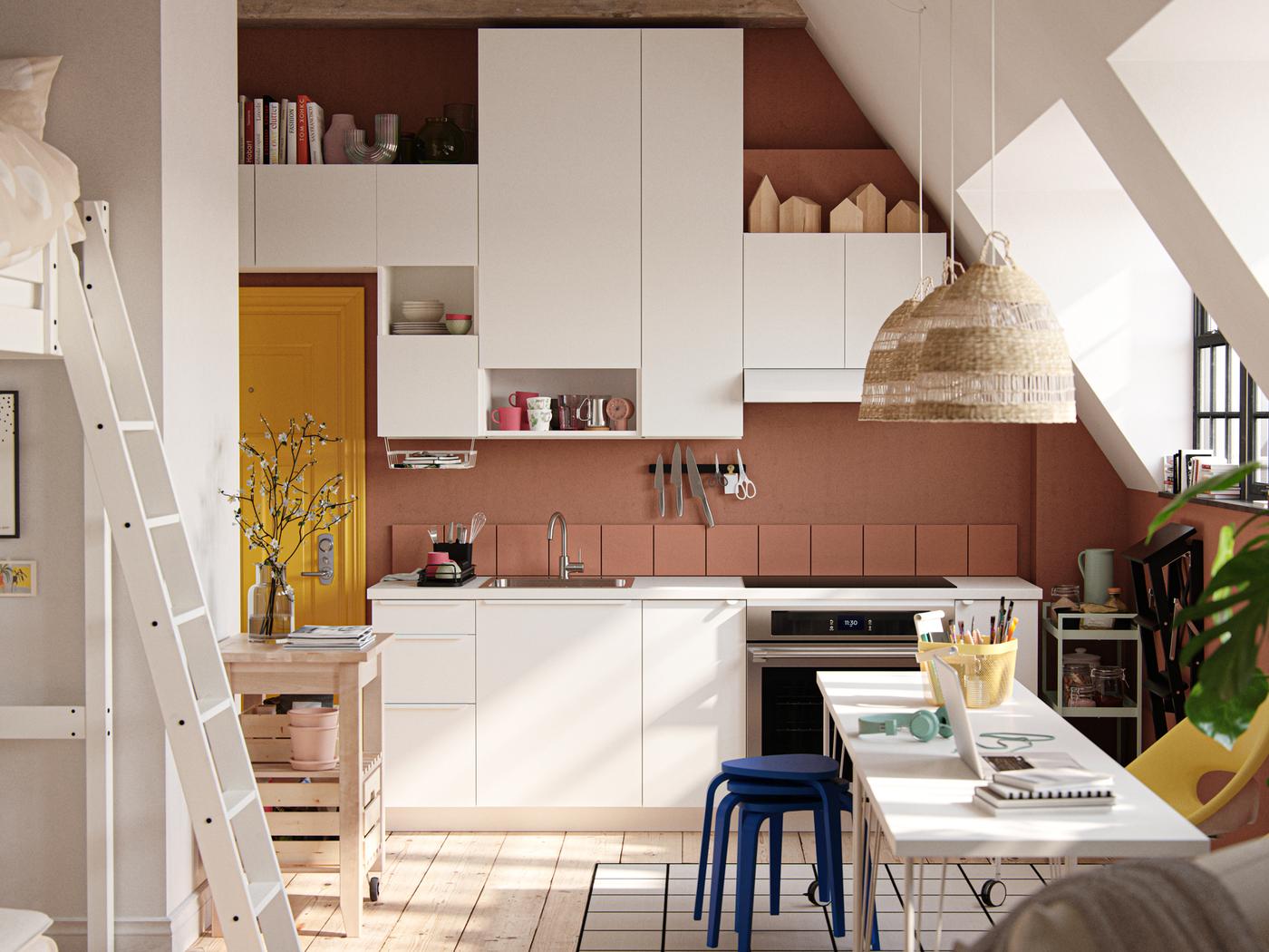 Un estudio con techo inclinado, ventanas de estilo industrial, paredes de color terracota oscuro y frentes de cocina VALLSTENA blancos.