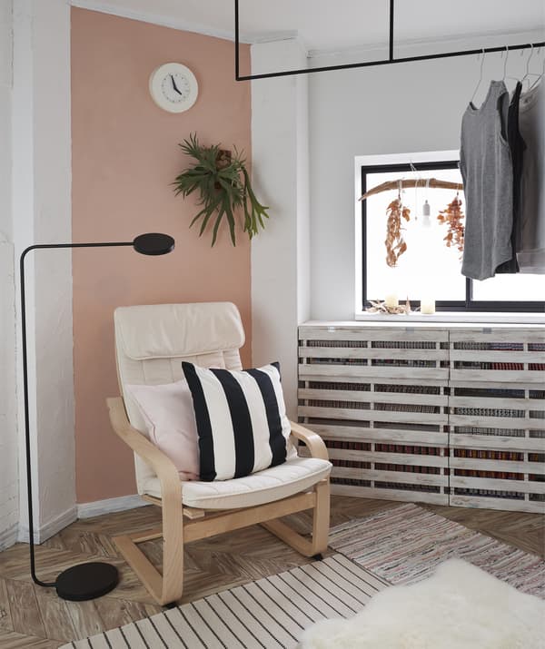 Un fauteuil et un lampadaire dans le coin d’une pièce avec un mur rose et des vêtements suspendus sur une tringle à la fenêtre.