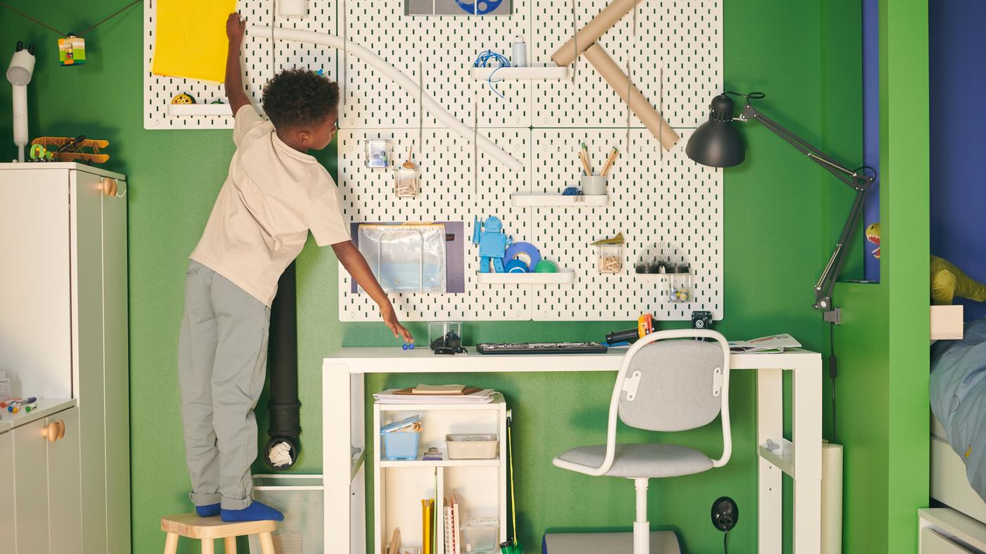 Un garçon debout sur un tabouret tend les bras vers une combinaison de panneaux perforés SKÅDIS accrochés au-dessus d’un bureau PÅHL blanc dans une chambre en vert et bleu.