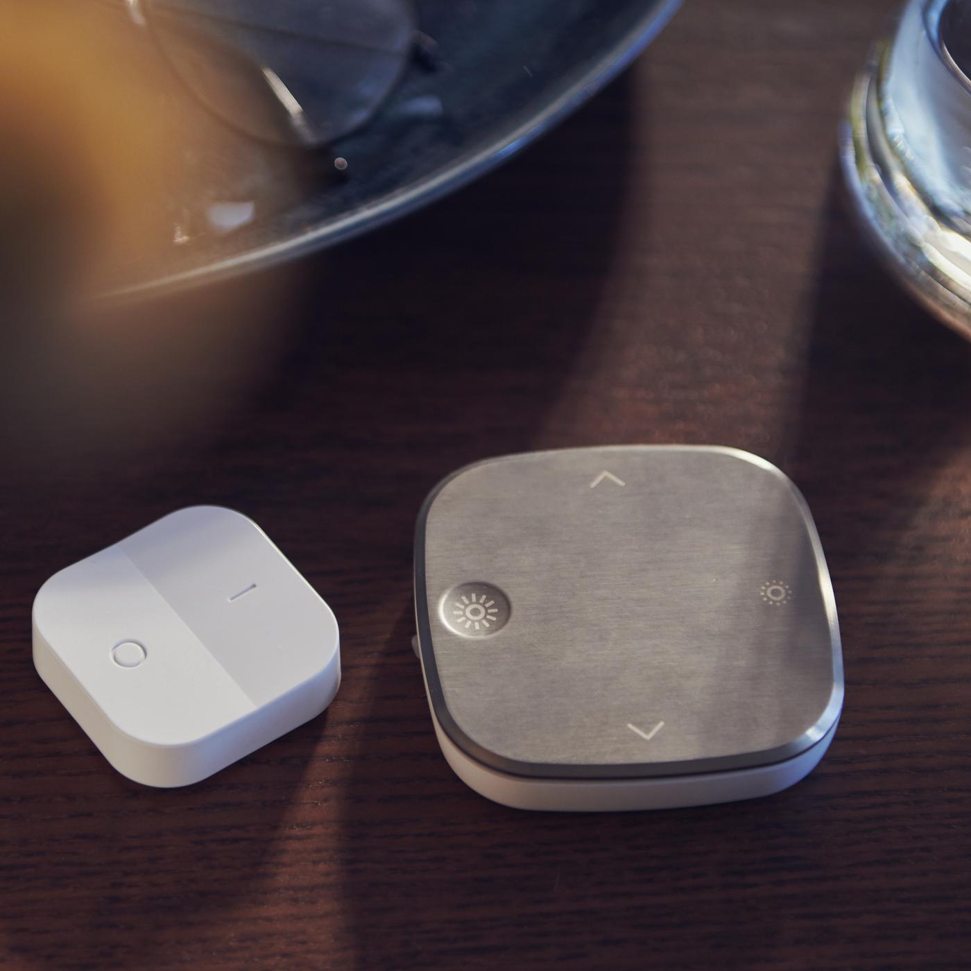 Un gradateur sans fil TRÅDFRI blanc et une télécommande STYRBAR couleur acier inoxydable sont posés sur le plateau d’une table en bois teintée foncée.