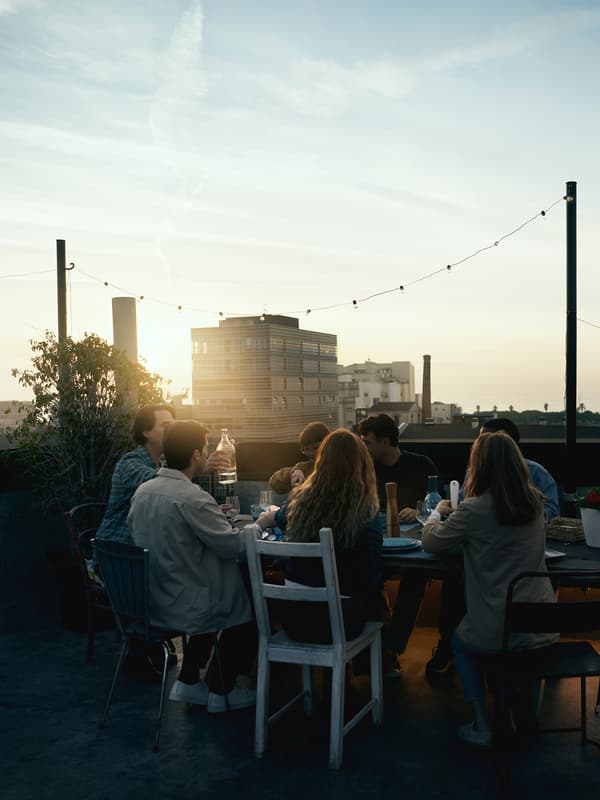 Un groupe de personnes assises sur des chaises autour d’une table au soleil couchant, en train de boire et manger, des buildings à l’arrière-plan.