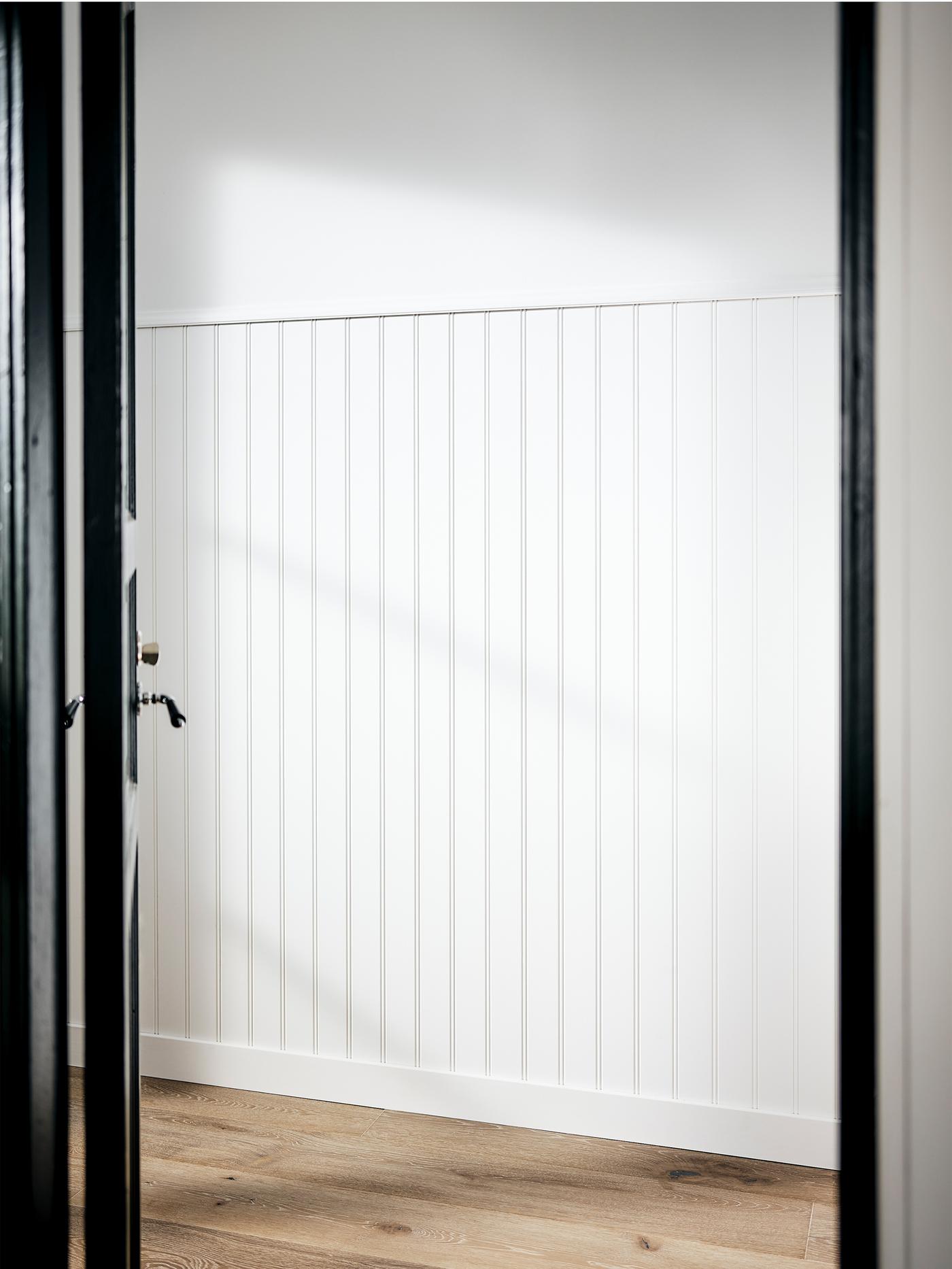 Un hall d’entrée au mur peint en blanc couvert jusqu’à mi-hauteur de panneaux en bois, et au sol en bois.