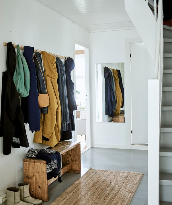 Un hall d’entrée blanc avec une rangée de crochets dans le style Shaker, un banc en bois, un tapis sisal au sol et un miroir accroché au mur.