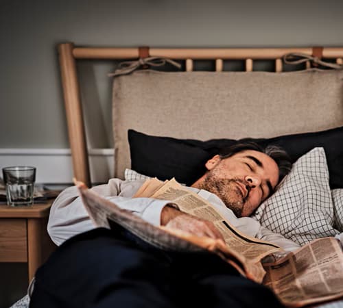 Un hombre acostado en una cama IKEA con dos almohadas y ropa de cama en tons azules. Al costado una mesita de noche con un vaso con agua.