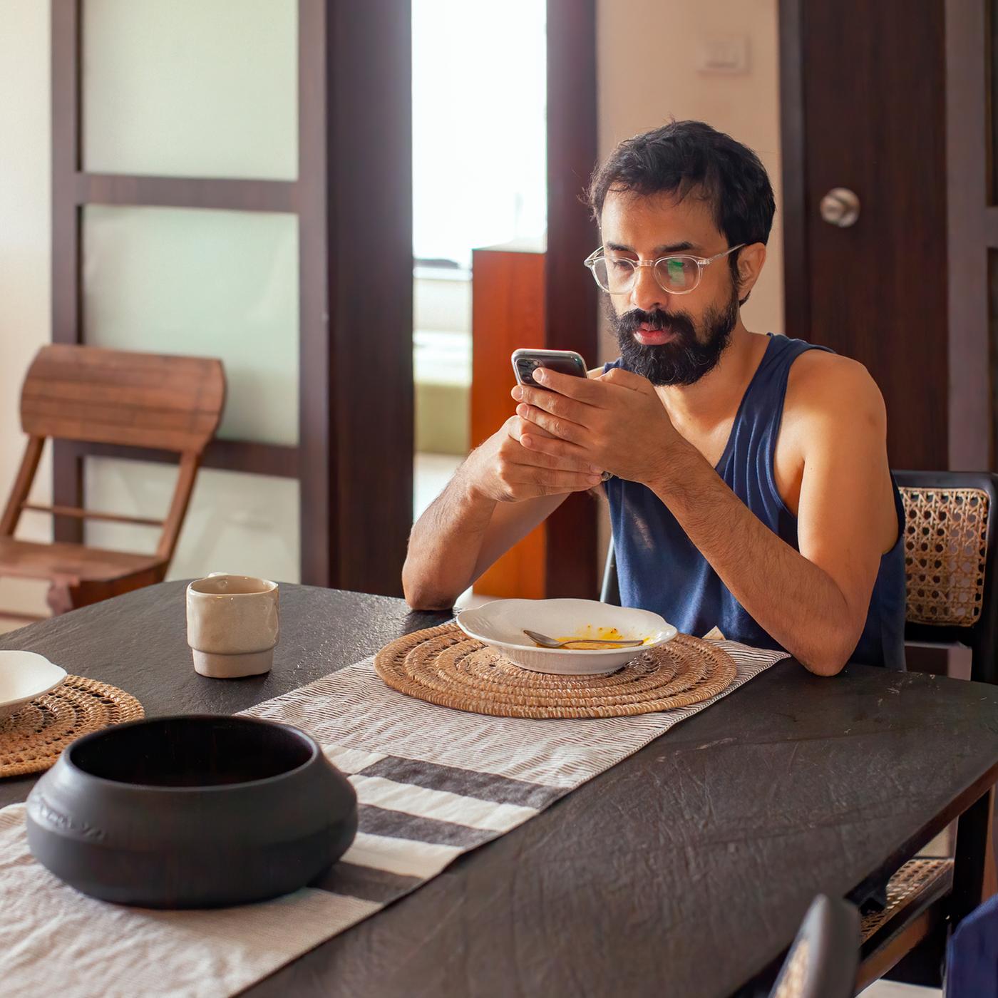 Un homme barbu aux cheveux foncés installé à table dans une cuisine vient de terminer de manger et regarde son téléphone.