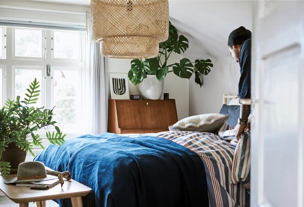 Un homme fait un lit avec une couette rayée rouge, blanche et bleue et un plaid bleu foncé dans une pièce avec un éclairage au plafond allumé.