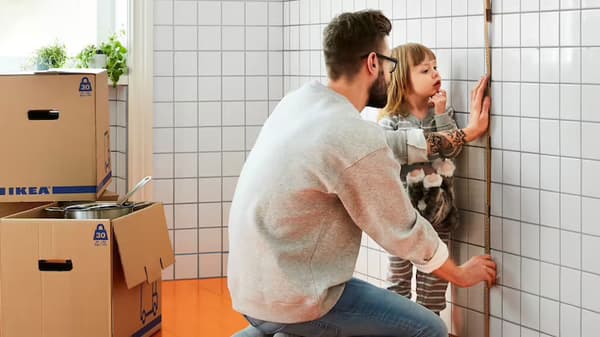 Un homme prend les mesures d’un mur alors que sa fille le regarde en arrière-plan. Des boîtes de déménagement sont empilées dans un coin.