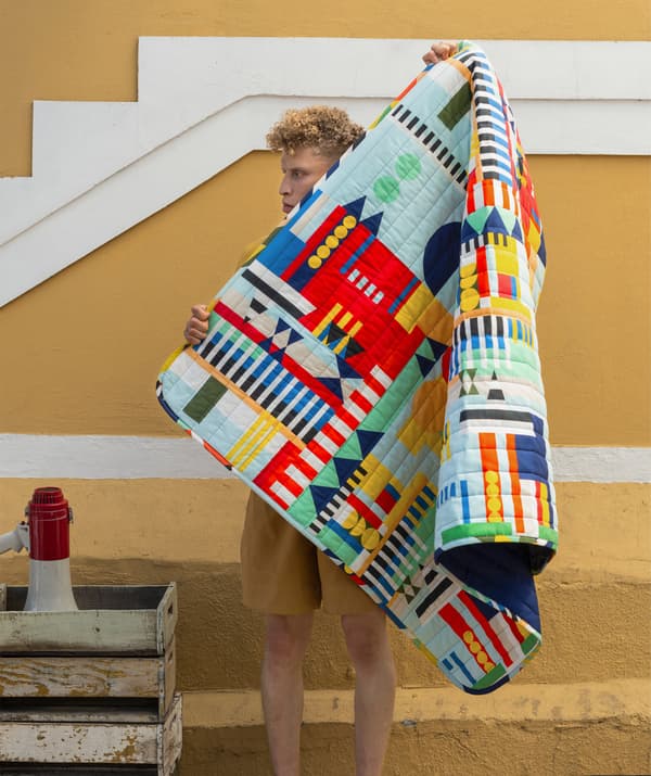 Un homme tenant une couverture aux motifs multicolores devant un mur jaune.