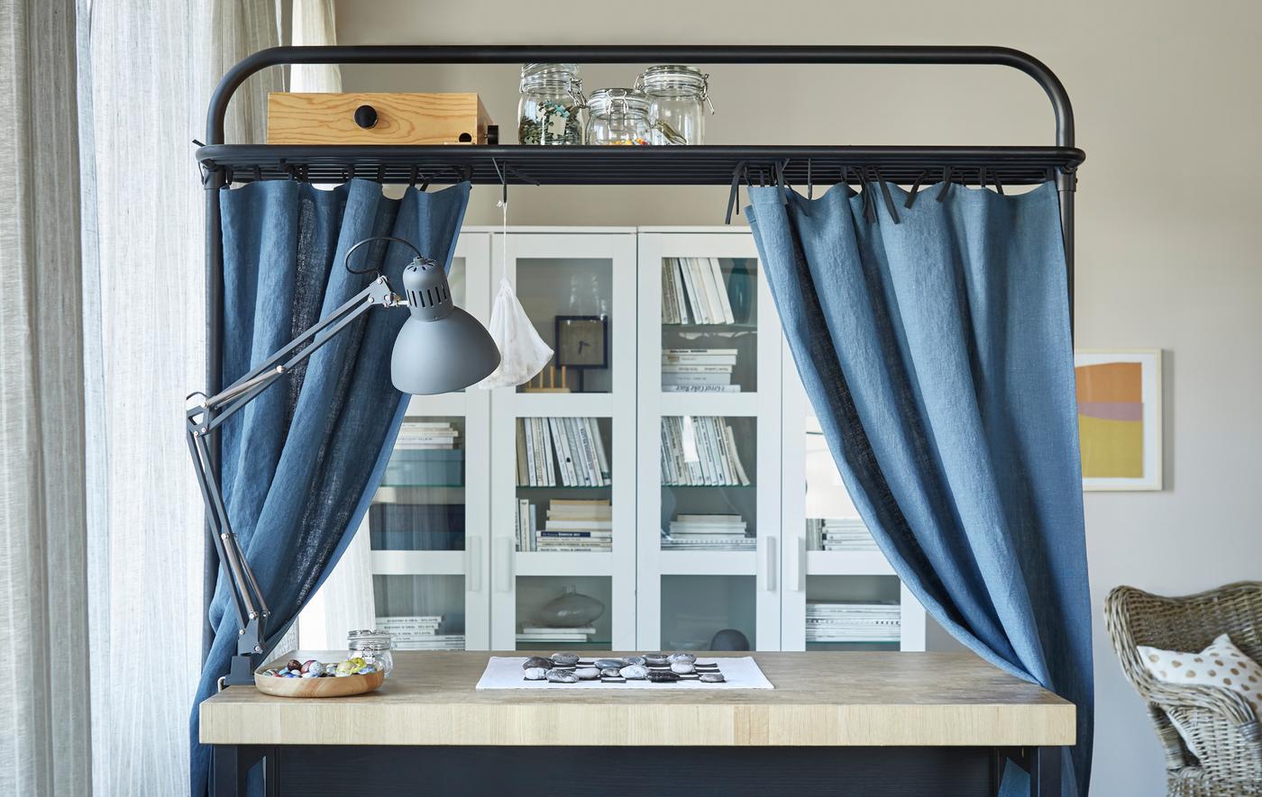 Un îlot de cuisine installé dans un séjour. Le support situé au-dessus, qui sert d’espace de rangement, est décoré de rideaux bleus.