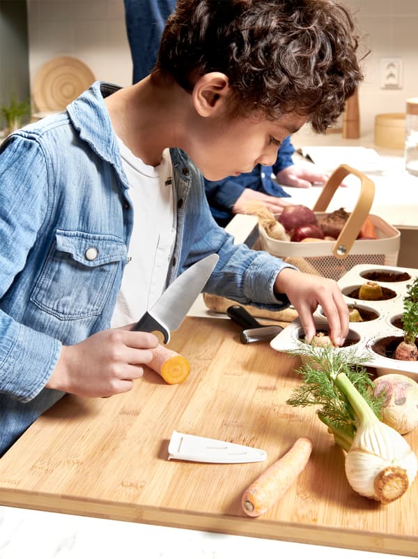 Un jeune garçon tenant un couteau plante la partie supérieure d’une carotte dans un moule à muffins plein de terreau, sur une surface en bois avec d’autres légumes.