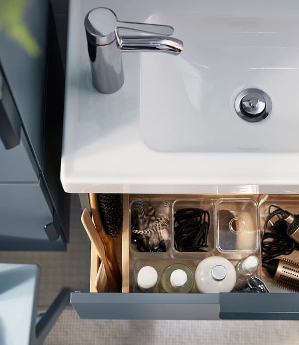 Un lavabo de salle de bains blanc vu d’en haut et un tiroir d’armoire bleu ouvert pour révéler des organiseurs en plastique transparent.