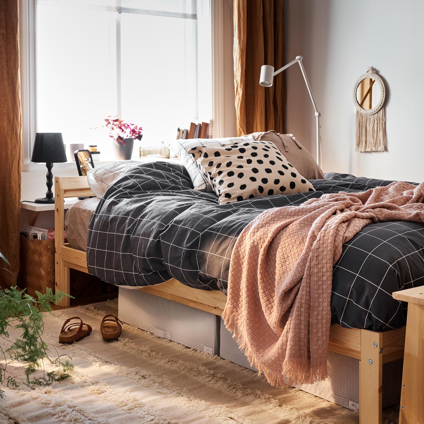 Un letto con struttura in pino e biancheria di colore bianco nero/a quadri, sotto al quale sono visibili alcune custodie bianche/grigie e un tappeto.