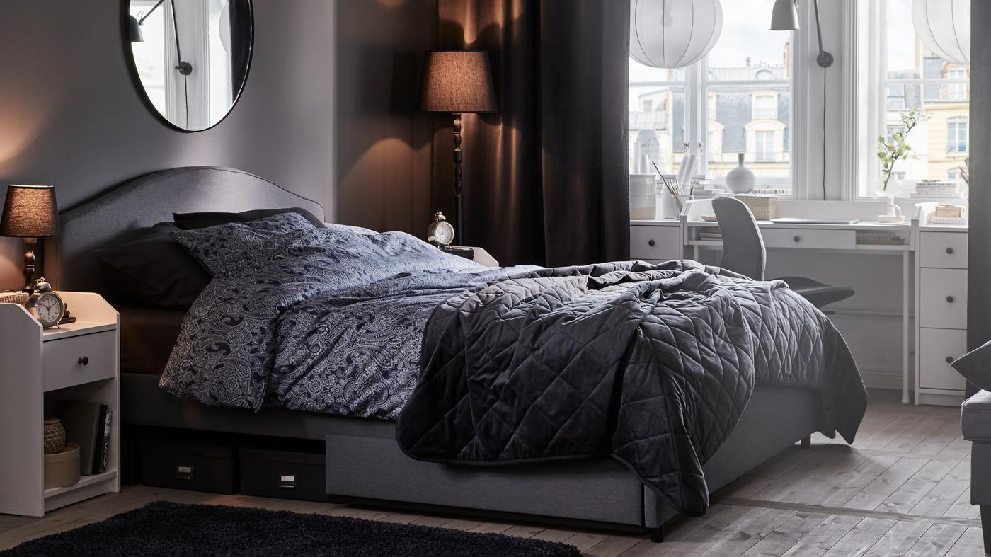 Un letto imbottito HAUGA grigio con biancheria da letto JÄTTEVALLMO e copriletto MJUKPLISTER contro la parete di una camera - IKEA.