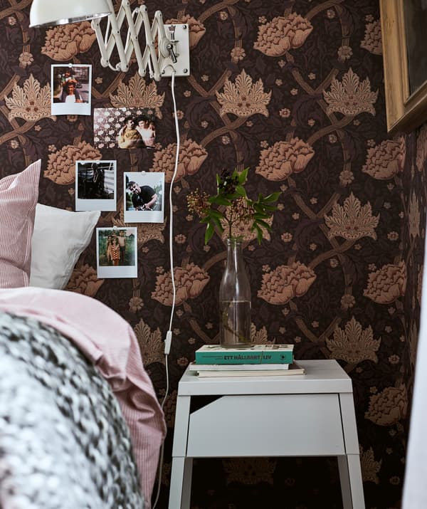Un lit avec du linge de lit rose à côté d’une table de nuit sur laquelle se trouvent des livres et une branche dans un vase, des polaroïds accrochés au mur.