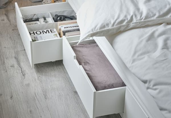 Un lit avec rangement NORDLI aux tiroirs ouverts, dans lesquels on peut voir du linge de lit plié ainsi que des livres, des magasines et des accessoires.