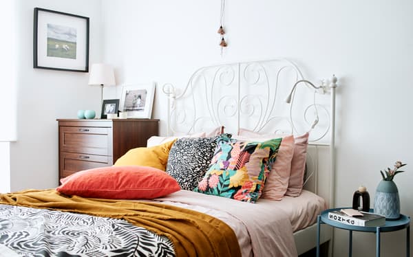 Un lit avec tête de lit blanche en fer forgé, une couette rose, un plaid jaune foncé et des coussins en rose, orange, doré et monochrome.