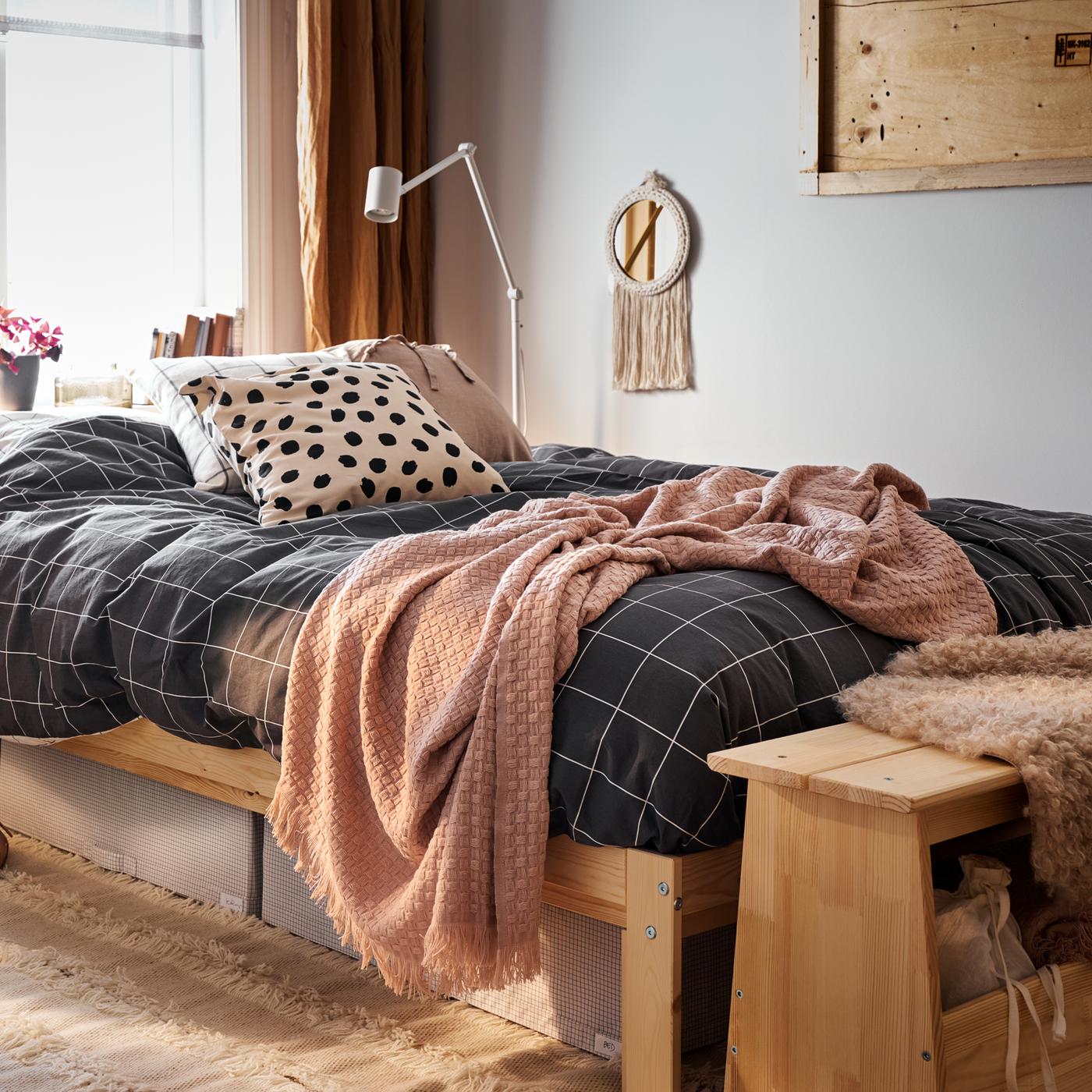 Un lit avec un cadre en pin et du linge de lit à carreaux noirs, plus des caisses de rangement blanches/grises et un tapis en dessous.