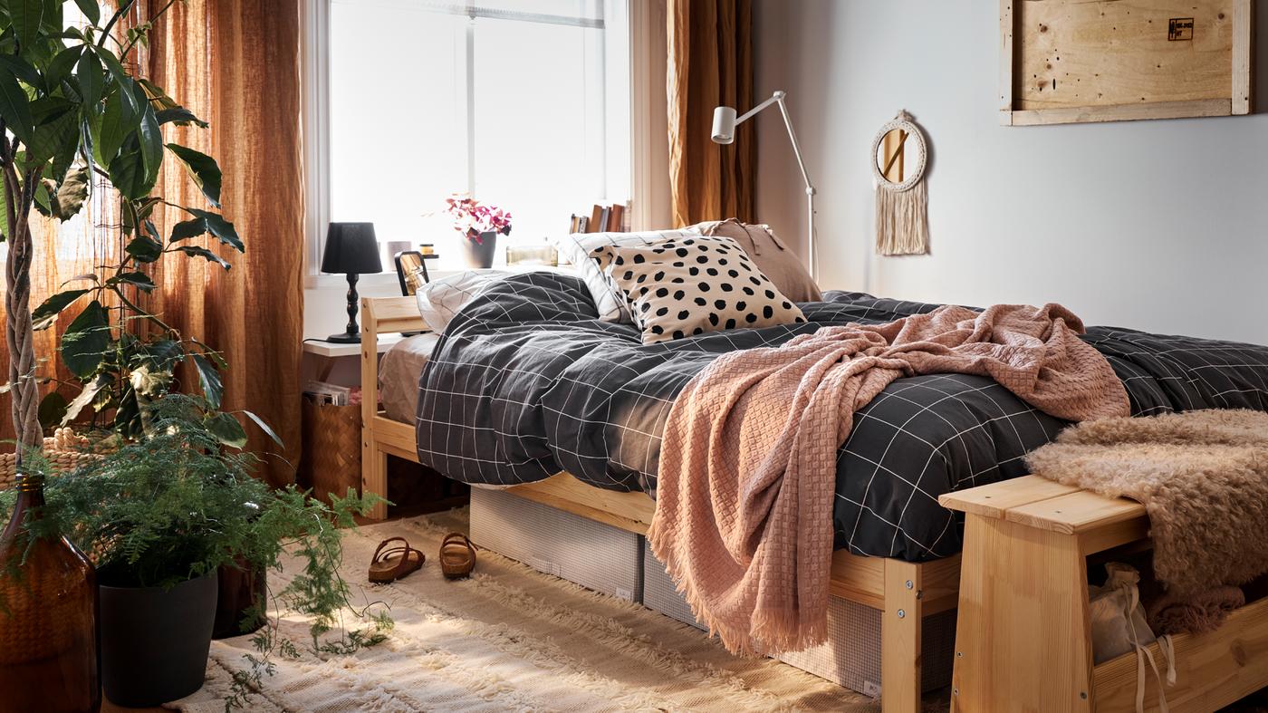 Un lit avec une structure de lit en pin recouvert de linge de lit à carreaux noirs et blancs, et des boîtes-tiroirs blanc-gris sur un tapis.