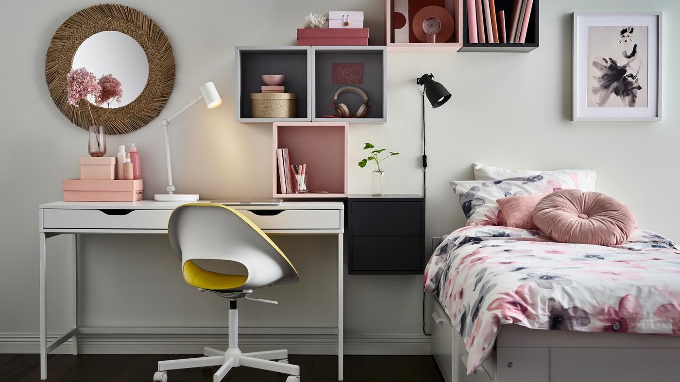 Un lit banquette BRIMNES blanc habillé de linge de lit LAPPNYCKLAR floral, à côté d’un bureau ALEX blanc en dessous d’étagères grises et roses.