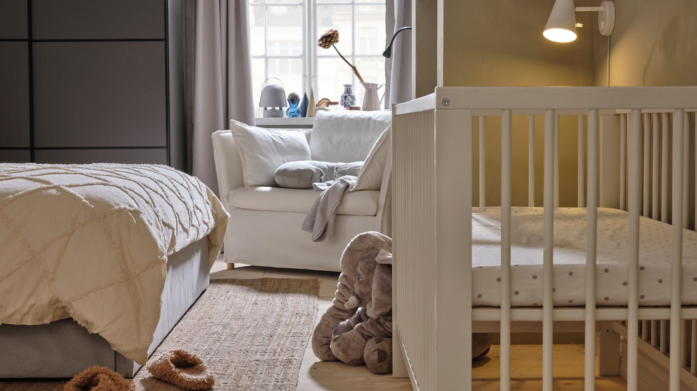 Un lit bébé GULLIVER blanc en face d’un lit de couleur grise, avec un fauteuil 1,5 place BACKSÄLEN blanc à l’arrière, près d’une fenêtre.