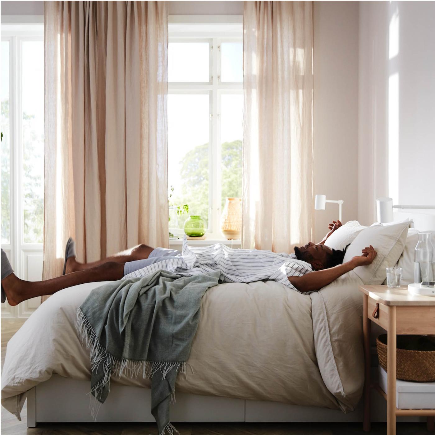 Un lit BJÖRKSNÄS en bouleau avec du linge de lit blanc et beige à côté d’une table de chevet blanche, une lampe de table en bambou et un purificateur d’air.
