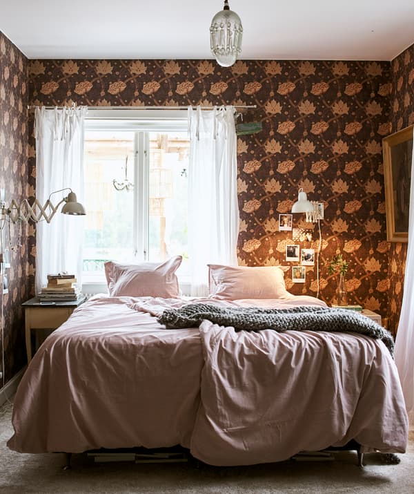 Un lit couvert de linge de lit rose et un plaid gris au centre d’une pièce avec un lustre et du papier peint à motif floral.