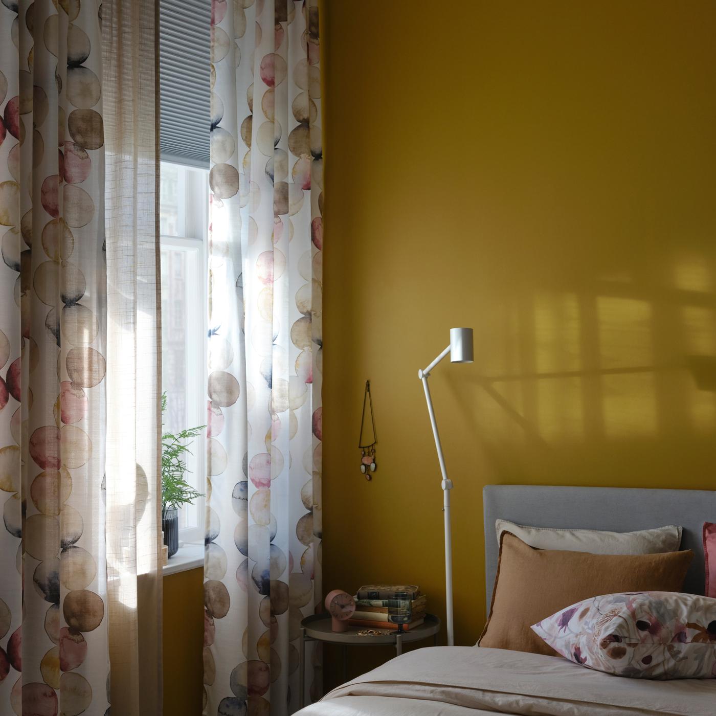 Un lit et un lampadaire/liseuse blanc près d’une fenêtre avec des rideaux SÄCKMAL multicolores et un store alvéolaire opaque.