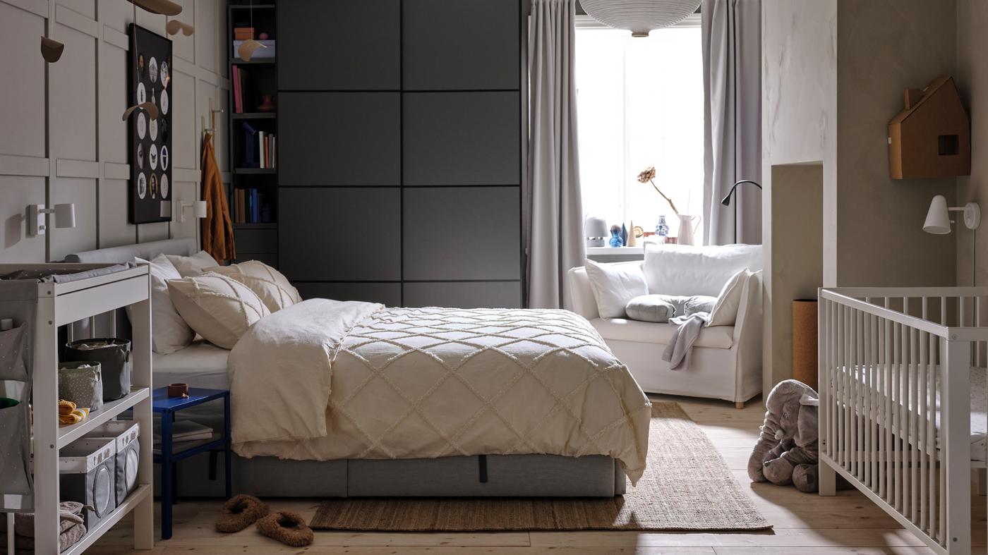 Un lit matelassé avec linge de lit NYPONLUGGMAL devant une armoire-penderie PAX/MEHAMN gris foncé et un large fauteuil blanc.