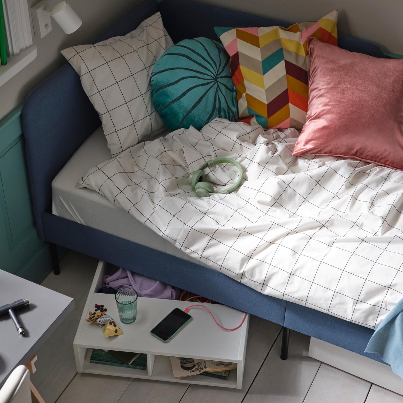 Un lit matelassé bleu avec du linge de lit VITKLÖVER à carreaux blancs et noirs, plusieurs coussins et un rangement sous le lit FREDVANG.
