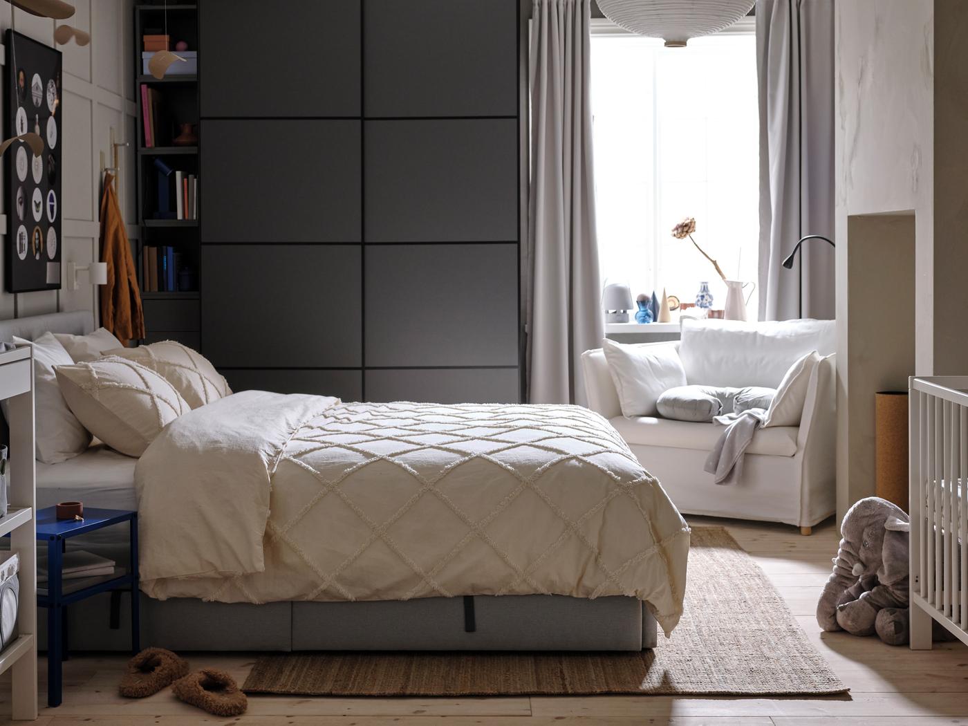Un lit matelassé gris avec linge de lit NYPONLUGGMAL devant une armoire-penderie PAX/MEHAMN gris foncé et un fauteuil blanc.