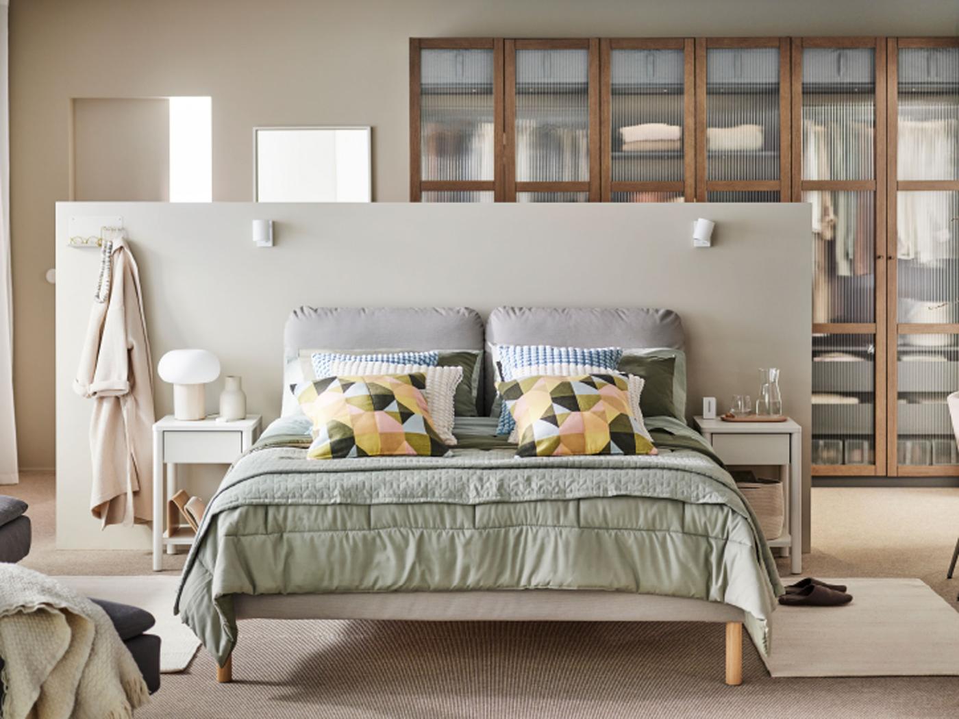 Un lit matelassé SAGESUND dans une chambre à coucher beige, avec un couvre-lit KÖLAX gris-vert et des coussins habillés de housses SVARTHÖ.