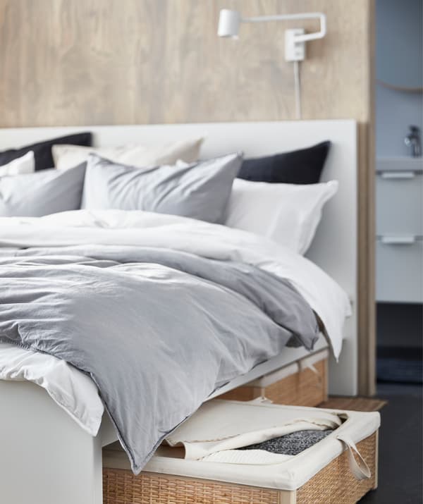 Un lit recouvert d’un linge de lit gris et blanc et sous lequel se trouve une boîte de rangement en rotin.