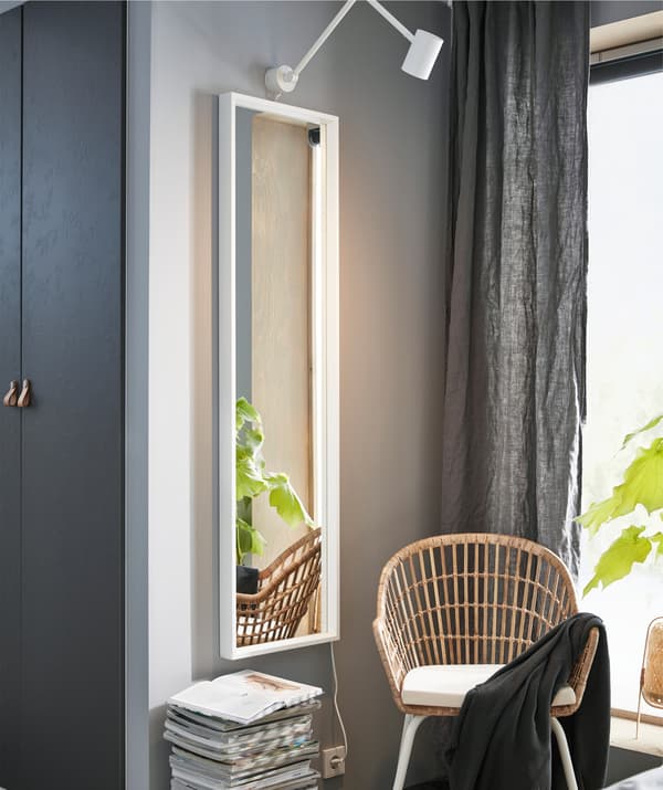 Un miroir en pied sur un mur gris, fauteuil en rotin et pile de magazines.