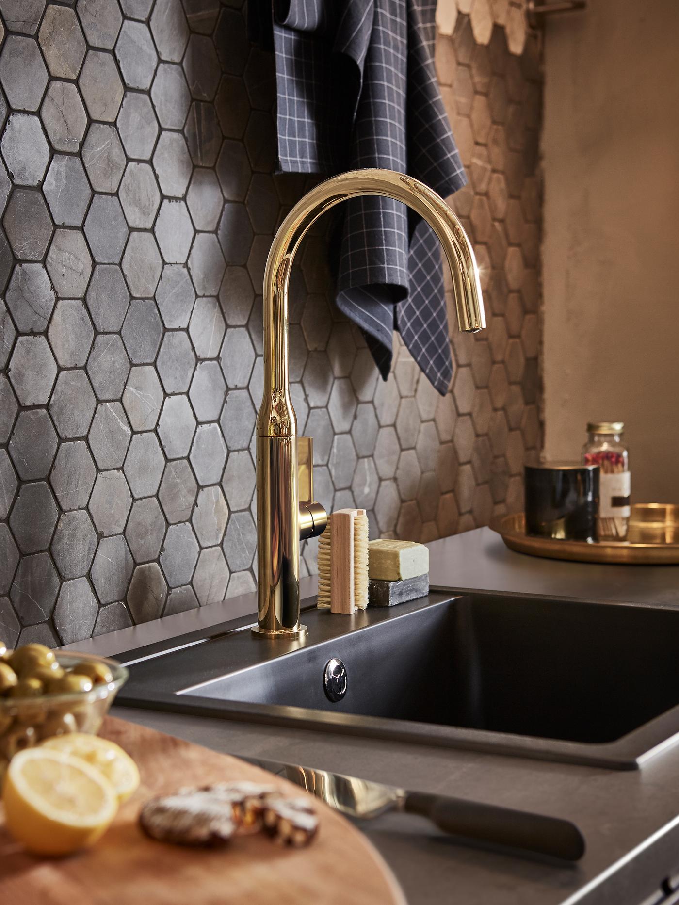 Un mitigeur couleur laiton et un évier de cuisine en composite de quartz noir devant un mur recouvert de carrelage hexagonal.